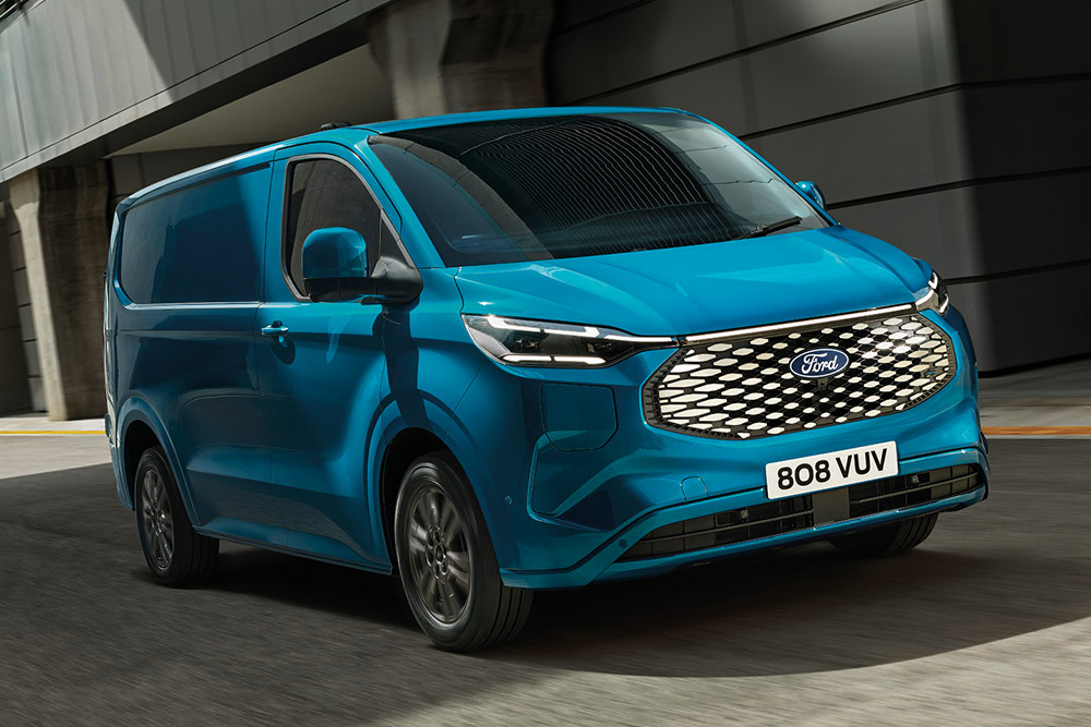 Niebieski Ford E-Transit Custom jadący drogą przy budynku magazynu.