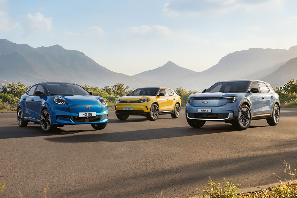 Ford Puma Gen-E, Capri oraz Explorer stoją na asfaltowej drodze na tle gór i zielonego krajobrazu w słoneczny dzień.