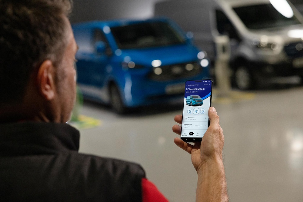 Mężczyzna trzyma smartfon z aplikacją Ford E-Transit Custom. W tle niebieski i biały van w warsztacie.