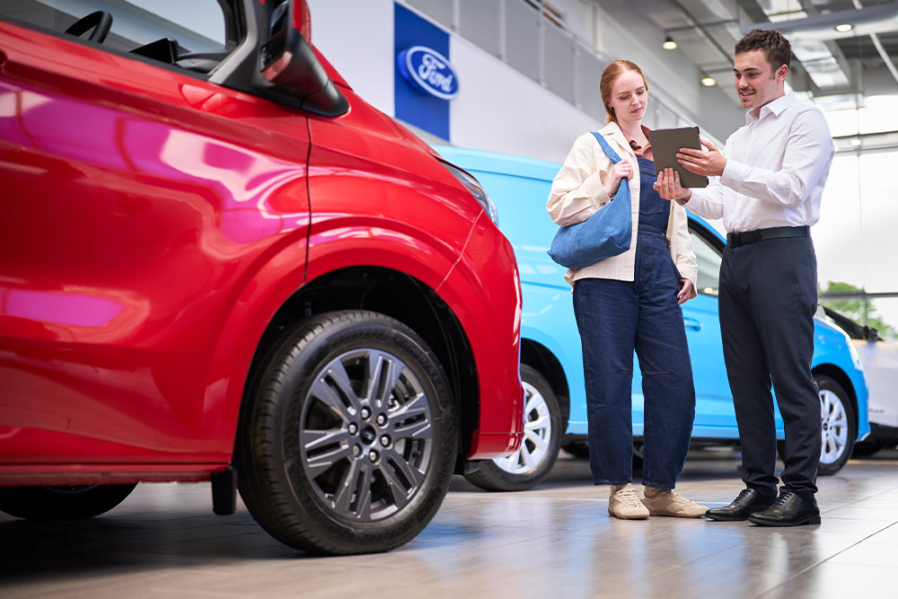 Two people in a Ford showroom looking at a tablet, with a red Ford van in the foreground and a blue Ford van behind.