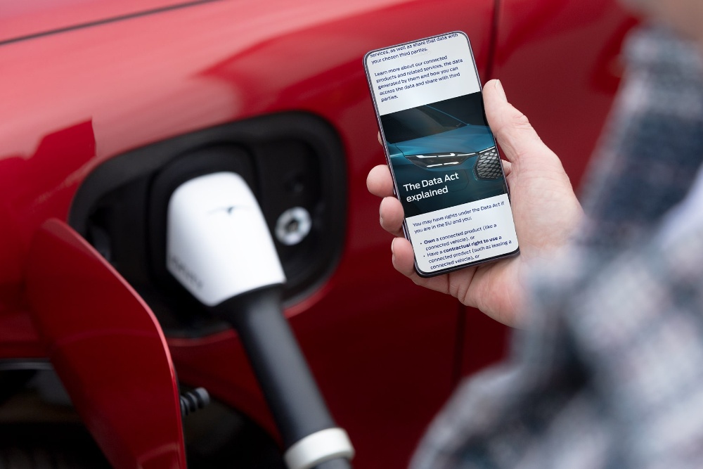 Osoba sprawdzająca smartfon, a w tle ładujący się czerwony samochód elektryczny.