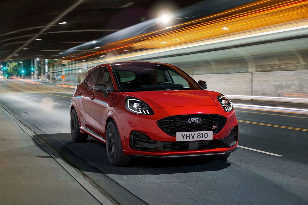 Czerwony sportowy SUV Ford Puma ST jadący jasno oświetloną miejską ulicą w nocy.
