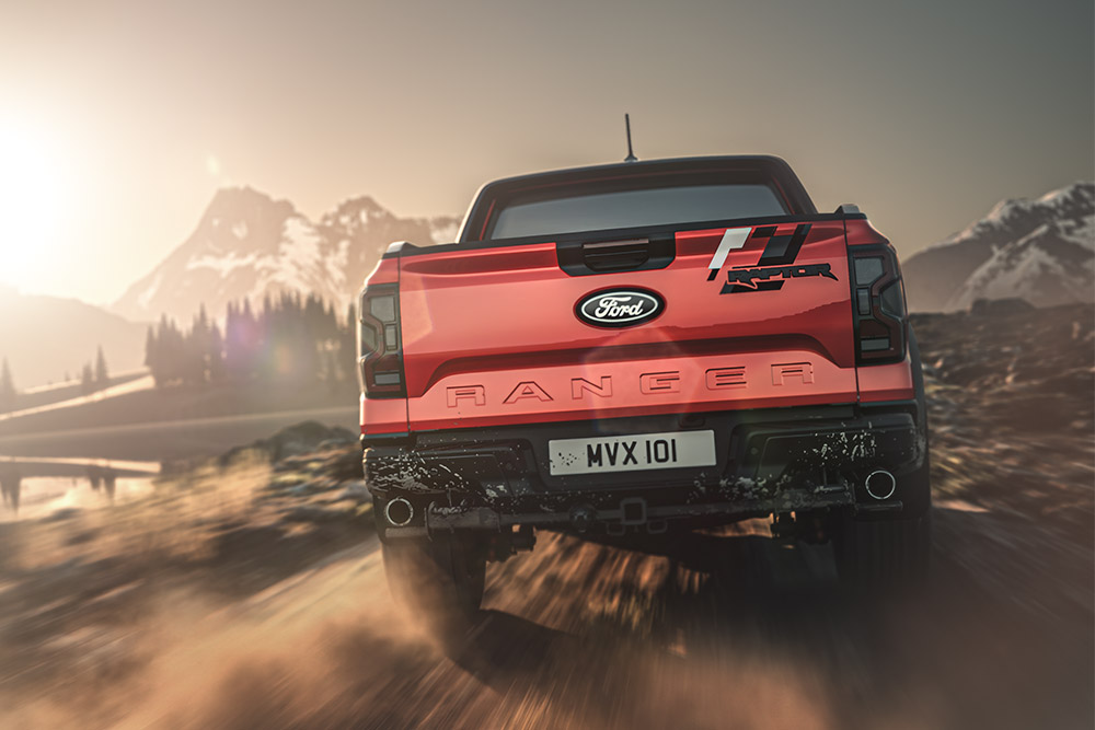 Widok z tyłu na czerwony samochód Ford Ranger Raptor poruszający się nieutwardzoną drogą z górami w tle.