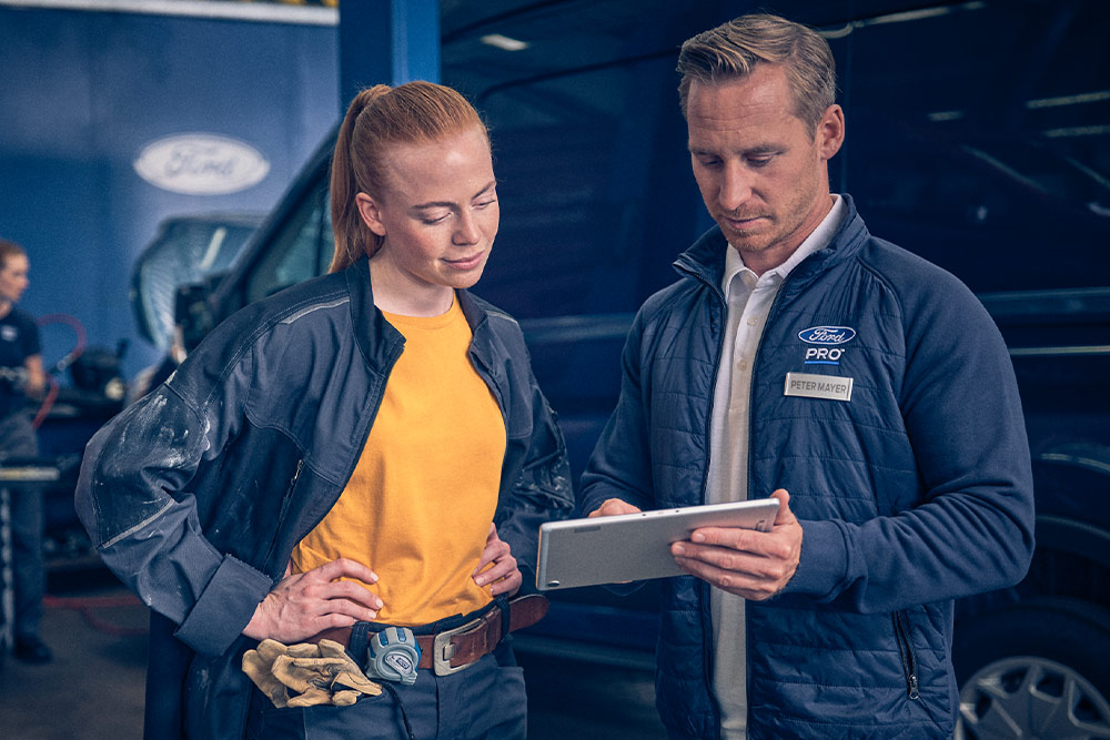 Dwóch mechaników w warsztacie patrzy na tablet przed vanem Ford E-Transit.