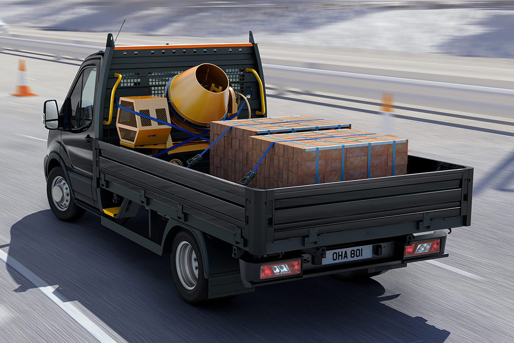 Rear 3/4 view of a Ford Transit Chassis Cab with a flatbed body, carrying a cement mixer and stacked bricks.