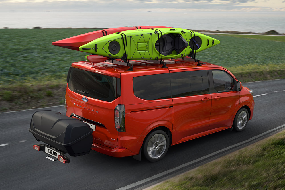 Czerwony Ford Pro Transit z dwoma kajakami na dachu, holujący bagażnik, jadący wzdłuż nadmorskiej drogi.