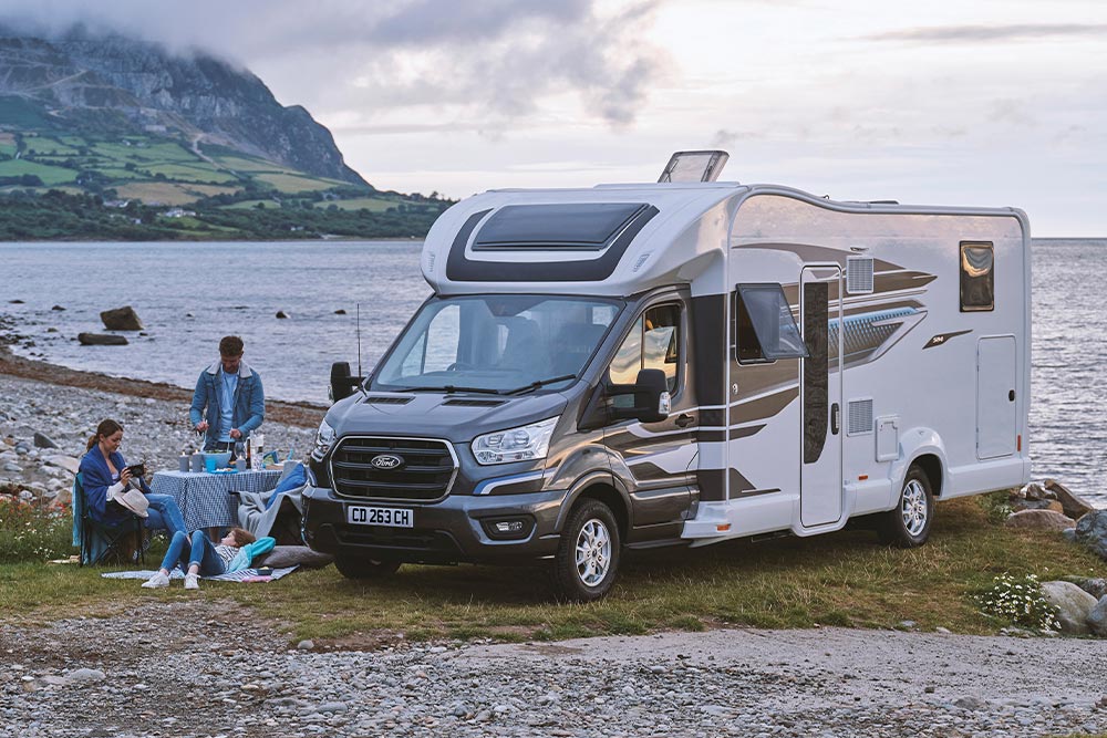  Para cieszy się napojami w krzesłach kempingowych obok szarego kampera Ford Transit Custom z podnoszonym dachem.