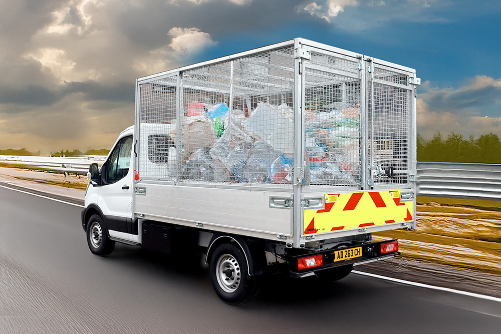 Widok z tyłu białej wywrotki klatkowej Ford Transit, pełnej śmieci, jadącej po drodze.