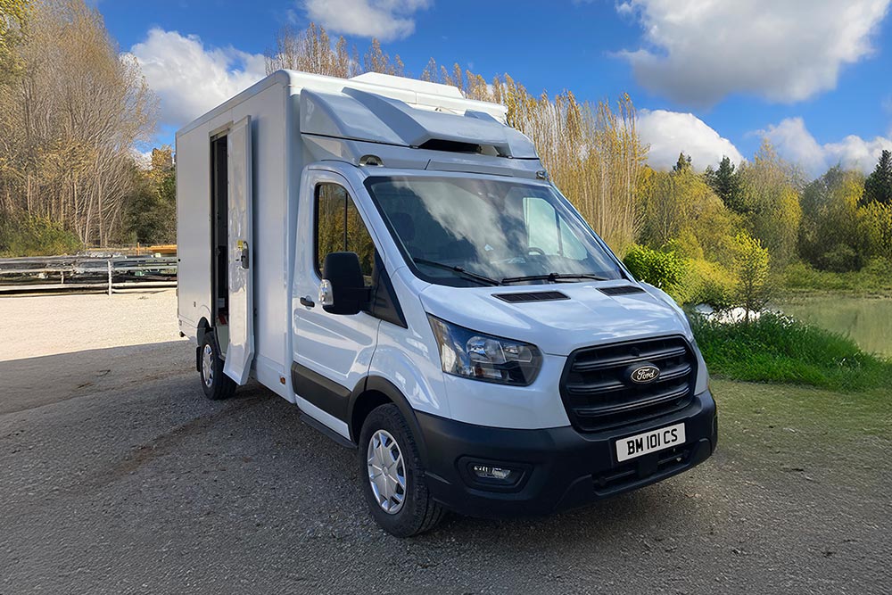 Widok 3/4 z przodu białego furgonu Ford Transit zaparkowanego nad jeziorem z drzewami.
