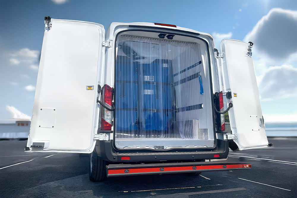 Widok z tyłu białego chłodniczego vana Ford Transit z otwartymi drzwiami i plastikowymi kurtynami paskowymi.